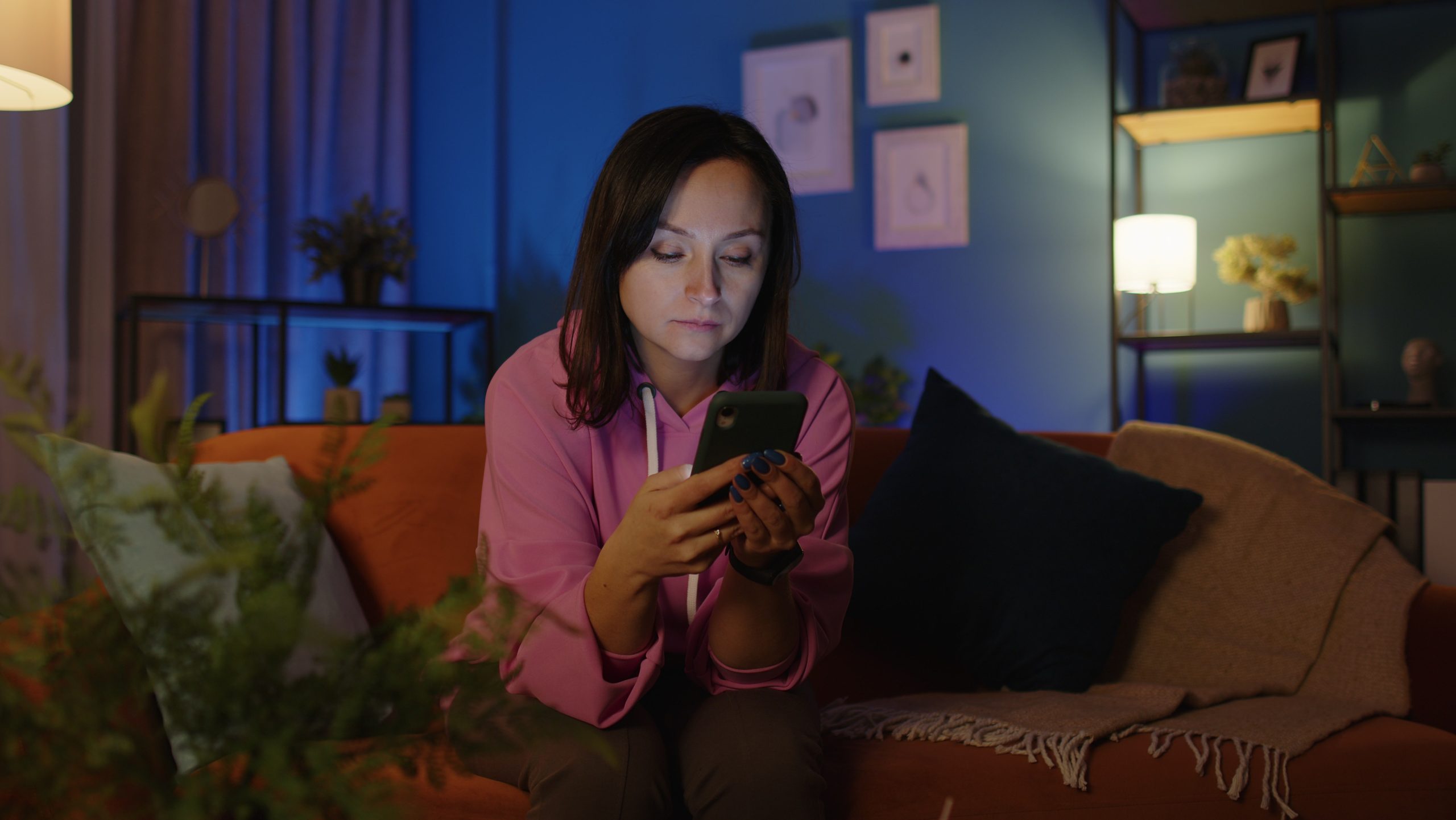 Woman texting message on smartphone, scrolling on mobile phone and sitting on sofa at home alone Smiled young Caucasian woman texting message on smartphone and sitting on sofa at home alone. Female tapping, scrolling on mobile phone social media in living room. Cheerful isolated girl online relax